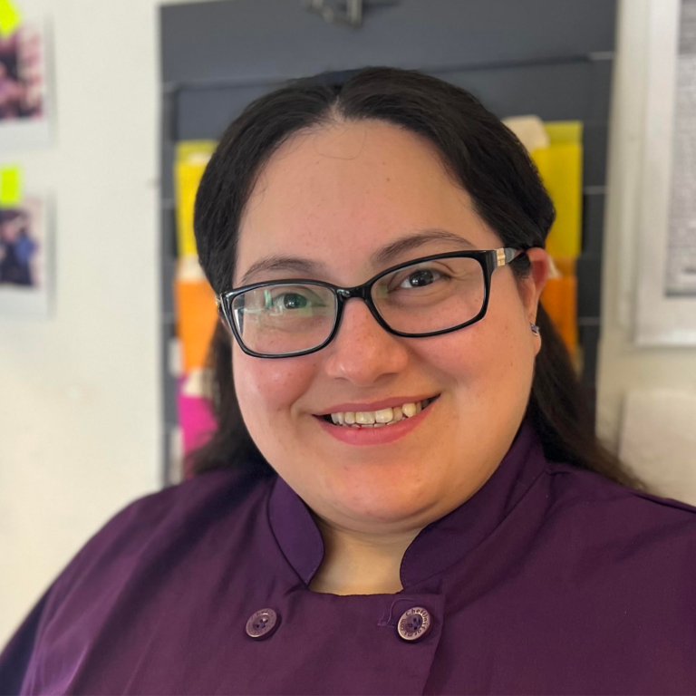 Smiling woman in a purple chef's jacket with glasses, standing in a colorful kitchen.