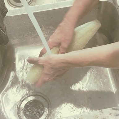 Hands washing a long vegetable under running water in a kitchen sink.